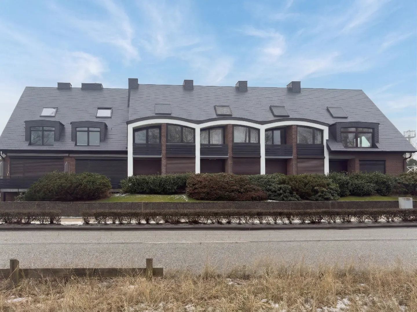 Row of brown brick townhouses with dark gray roofs and white trim, viewed from across a road.