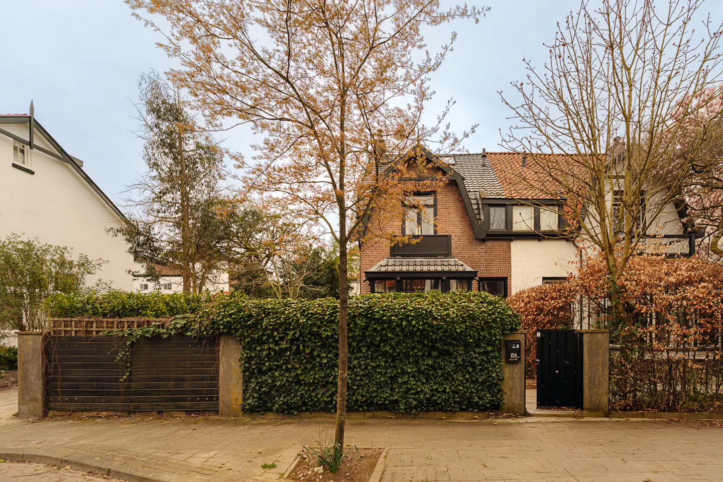 A two-story brick house with a dark roof and a black door is surrounded by green hedges and trees.