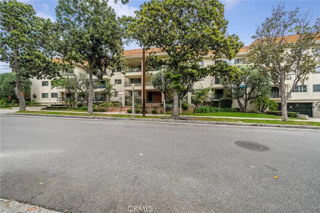 A three-story apartment building with balconies and a red tile roof, viewed from the street. Trees line the sidewalk.