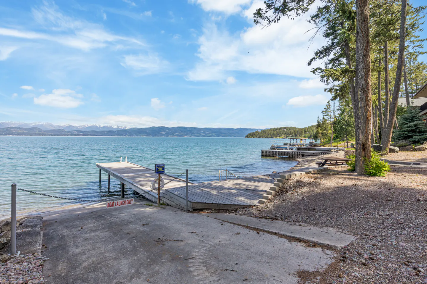 A scenic view of a boat launch with a wooden dock extending into a blue lake under a partly cloudy sky. Mountains are visible in the distance.