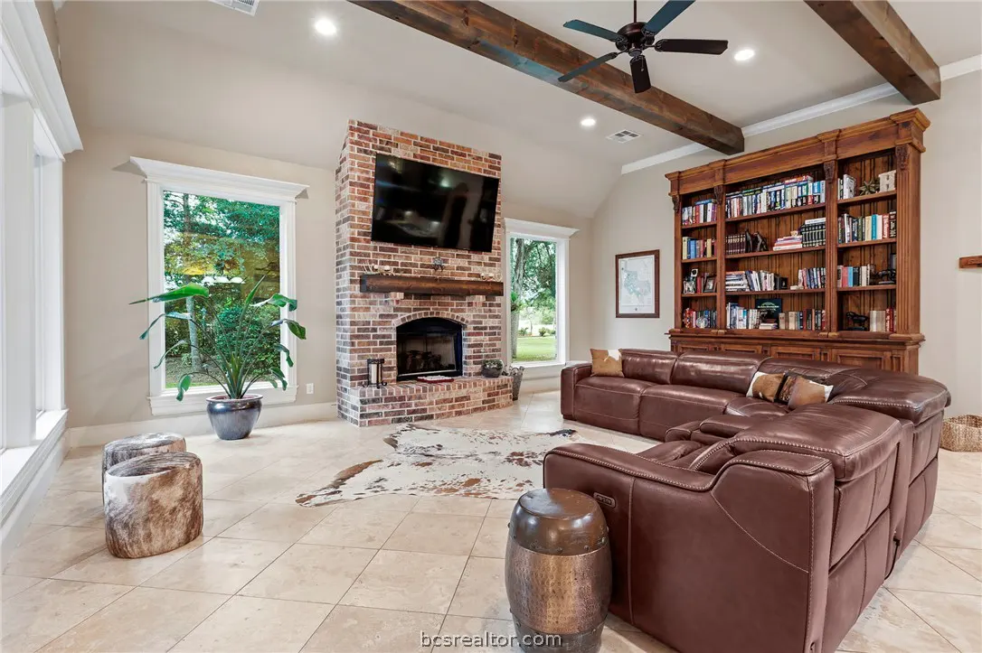 Living room with a brick fireplace, TV, brown leather sectional, and a large wooden bookcase. Windows overlook a green yard.