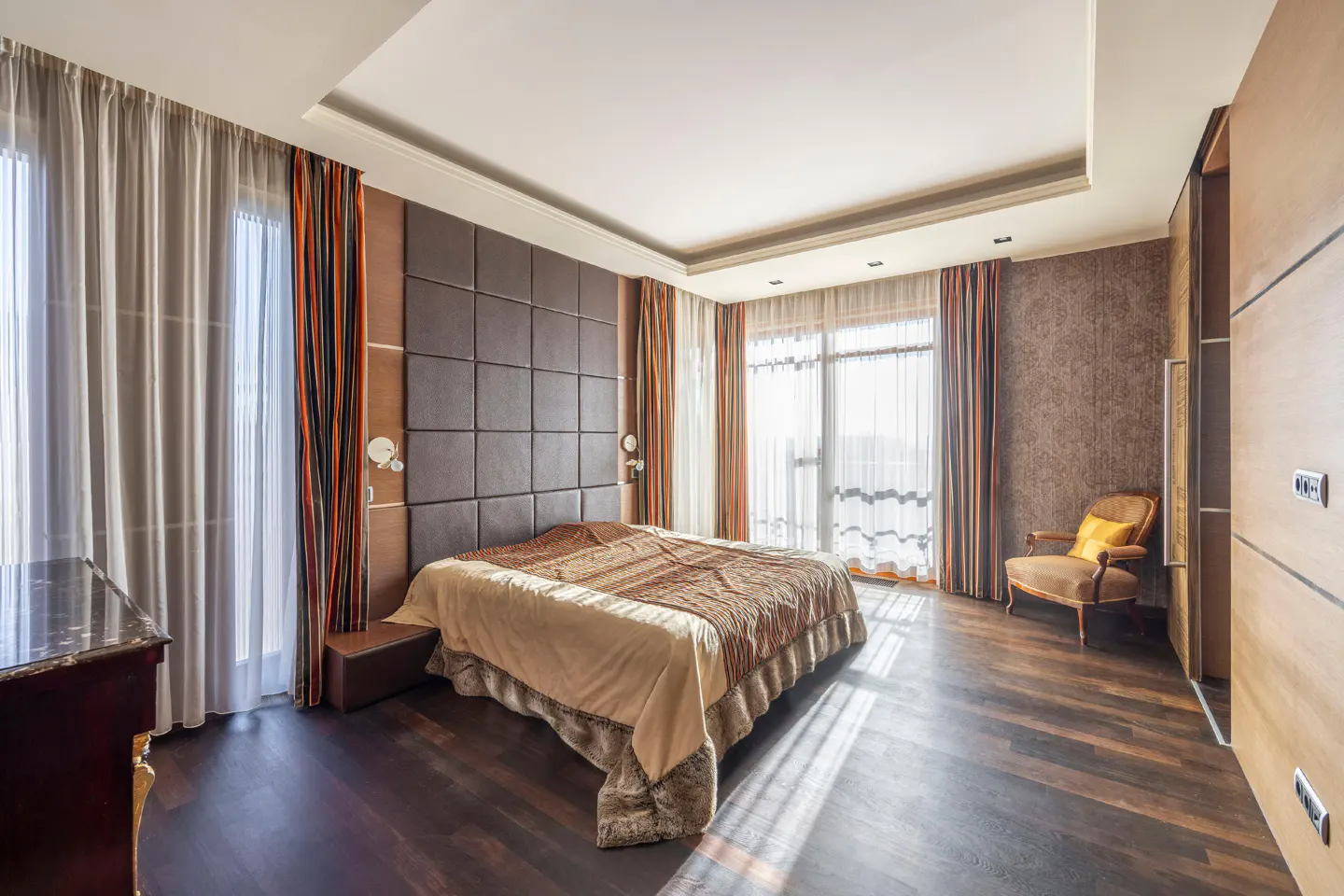 Bedroom with dark wood floors, a large bed with a brown headboard, and sheer curtains with orange stripes.