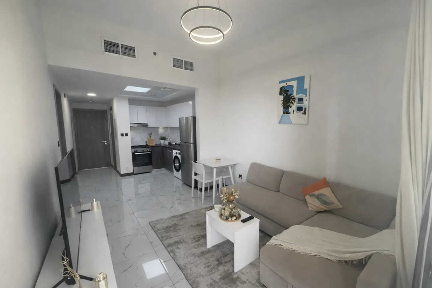 A bright, modern apartment interior with a gray sofa, white table, and kitchen in the background. A painting hangs on the white wall.