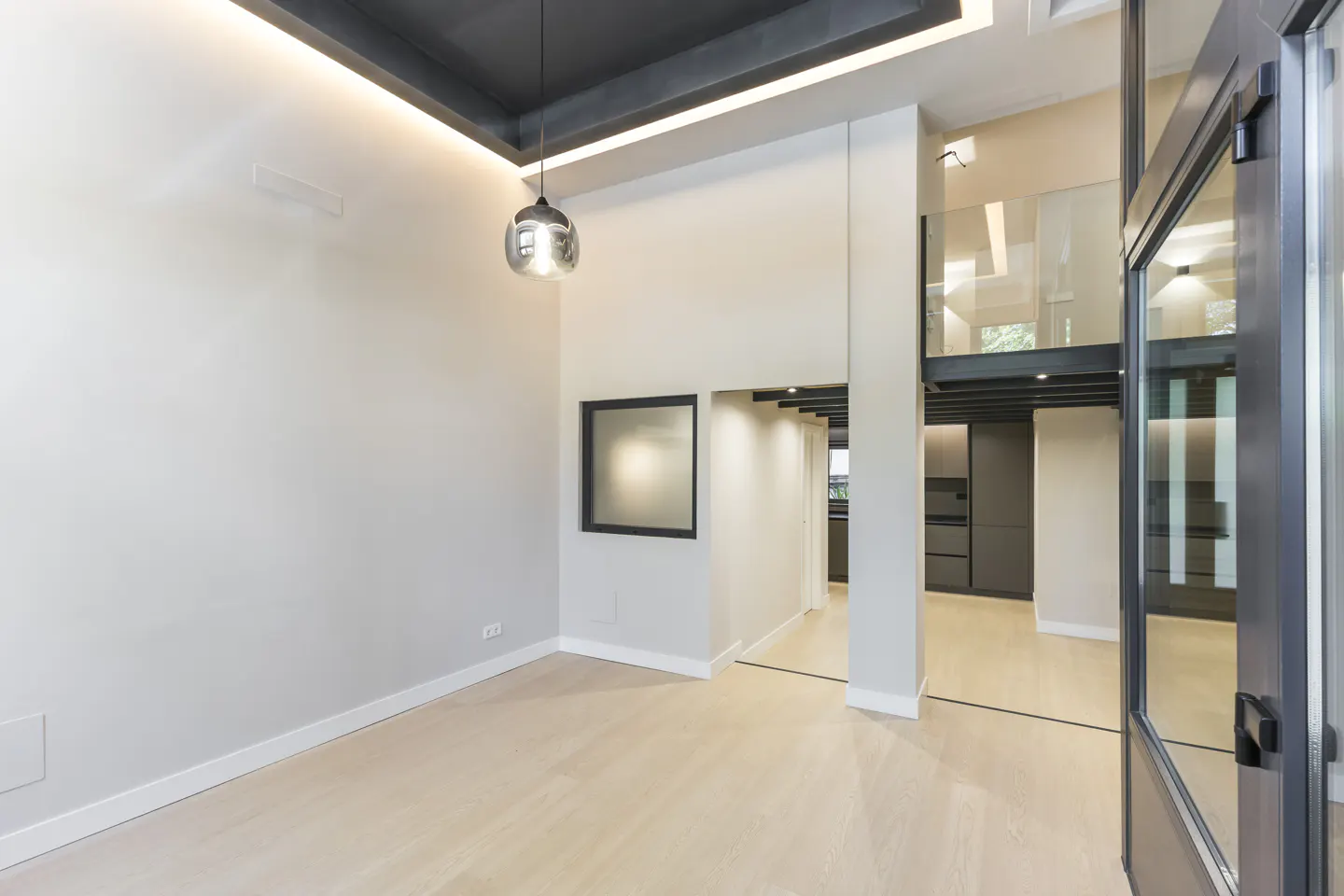 Modern living room with light wood floors, high ceilings, a pendant light, and a view into a kitchen with a loft level.