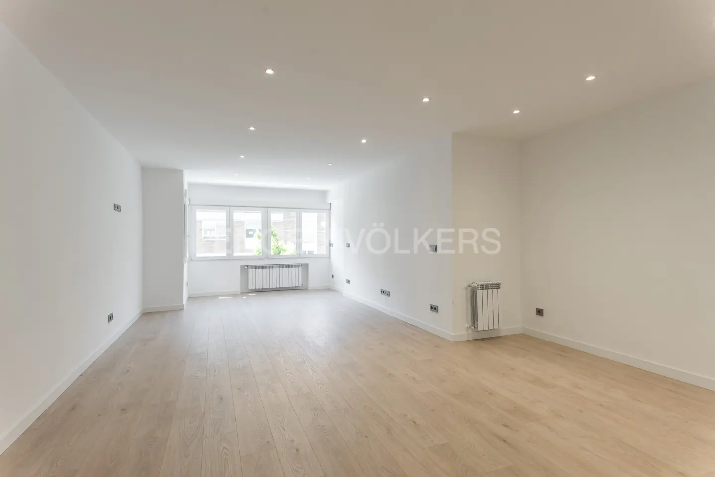 Bright, empty room with light wood floors, white walls, and recessed lighting. A window and radiator are visible.