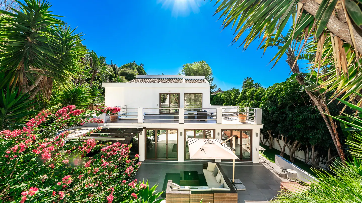 Two-story white house with a patio, surrounded by lush green trees and pink flowers under a bright blue sky.