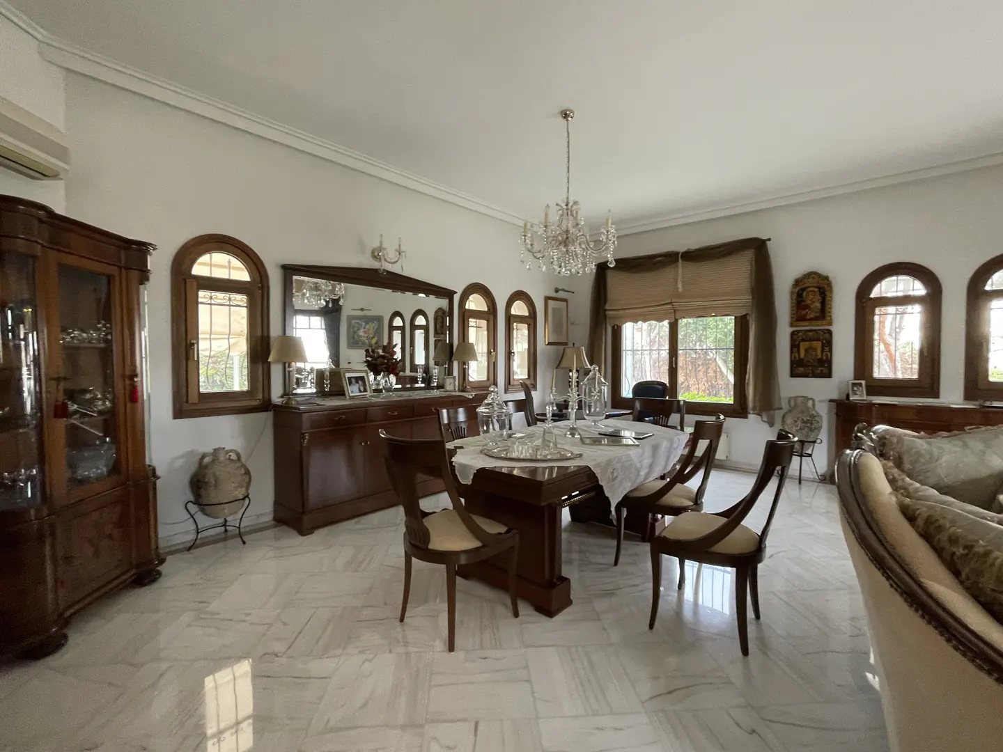 Formal dining room with dark wood furniture, white marble floors, a crystal chandelier, and arched windows.