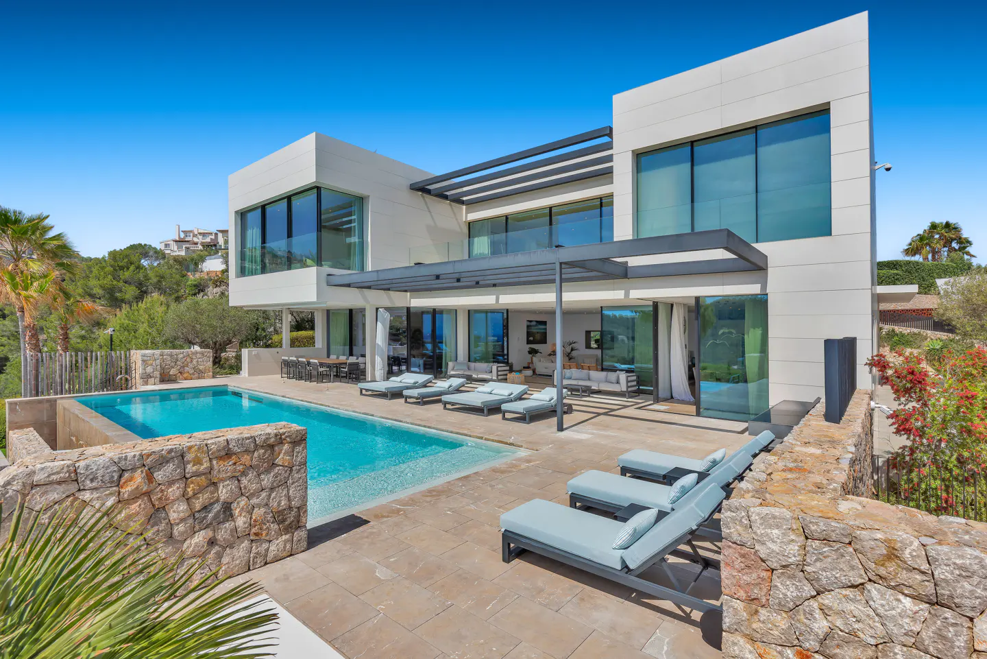 Modern white villa with a blue pool and lounge chairs on a stone patio under a clear blue sky.