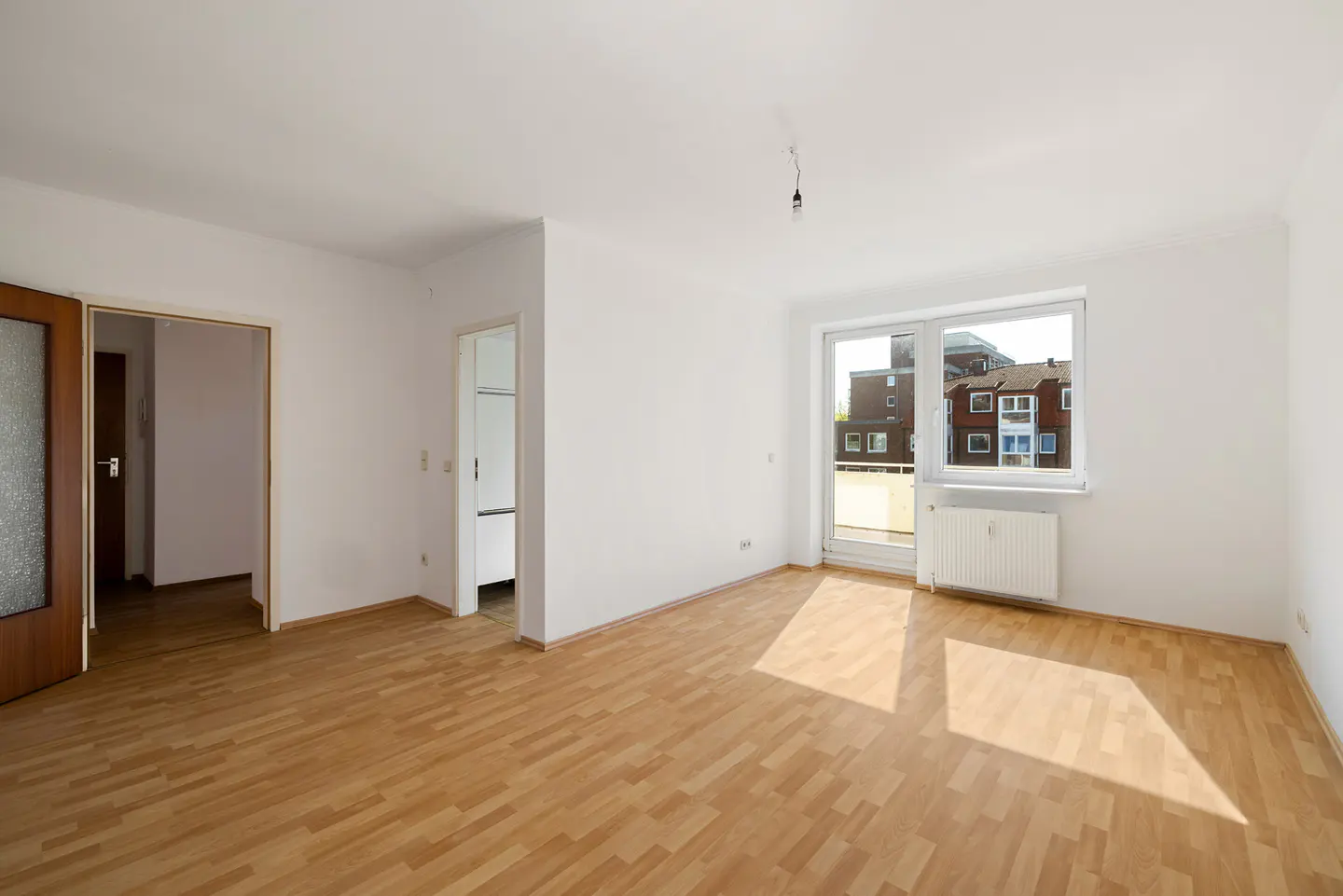 Bright, empty room with wood floors, white walls, and a large window showing a building outside.