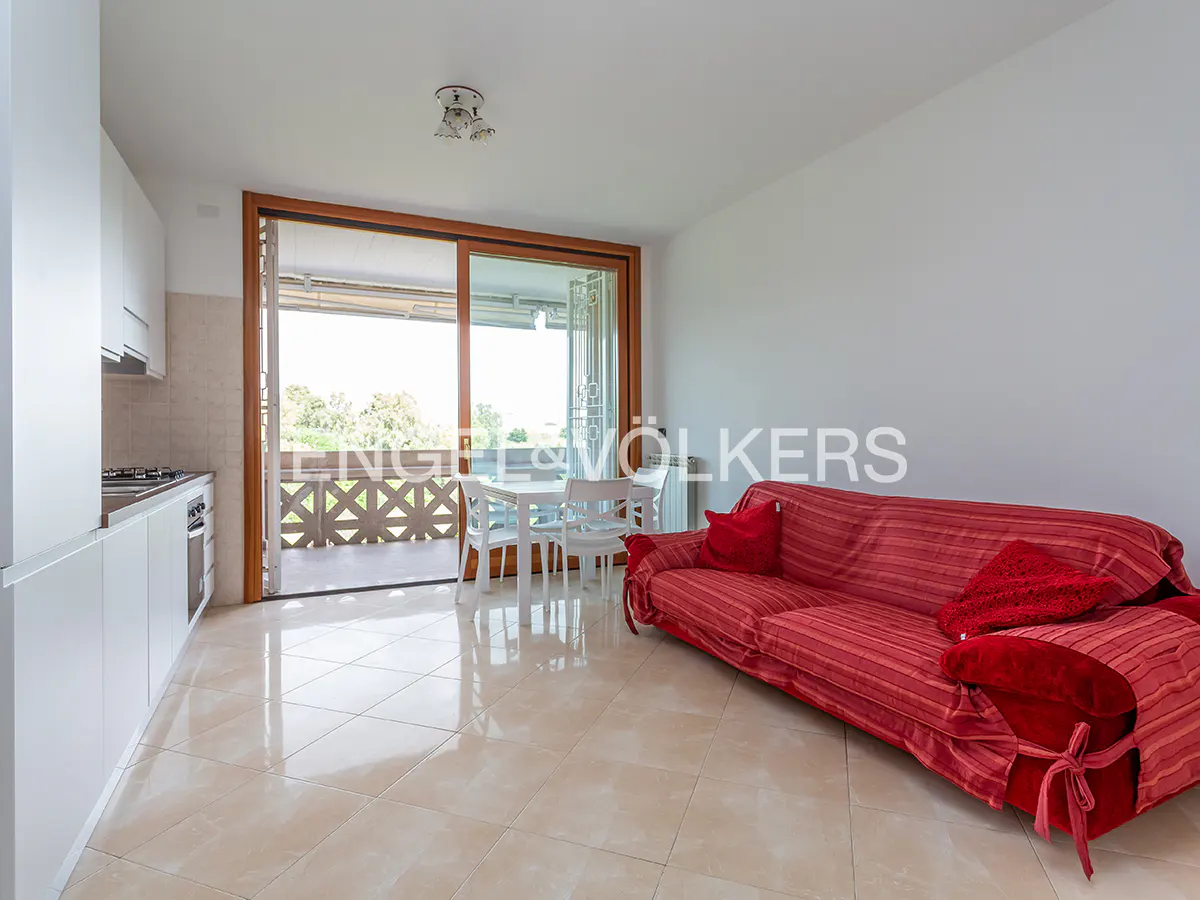 Bright apartment interior with white kitchen cabinets, a red sofa, and a dining table near a sliding glass door to a balcony.