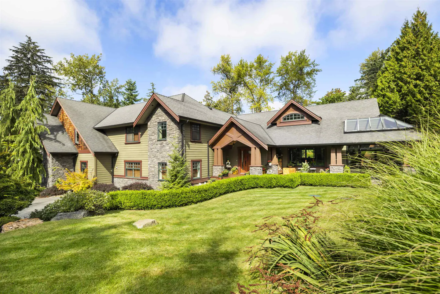 A large, two-story house with green siding, stone accents, and a gray roof sits on a lush green lawn. Trees surround the property.