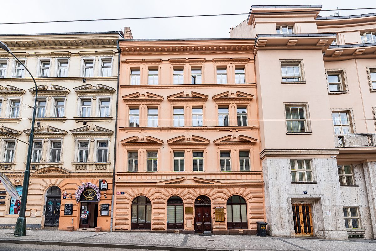 Street view of three buildings with ornate facades in Prague. The buildings are beige, peach, and cream colored.