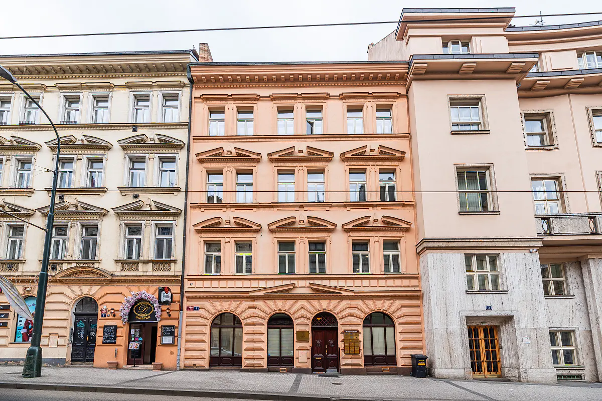 Street view of three buildings with ornate facades in Prague. The buildings are beige, peach, and cream colored.