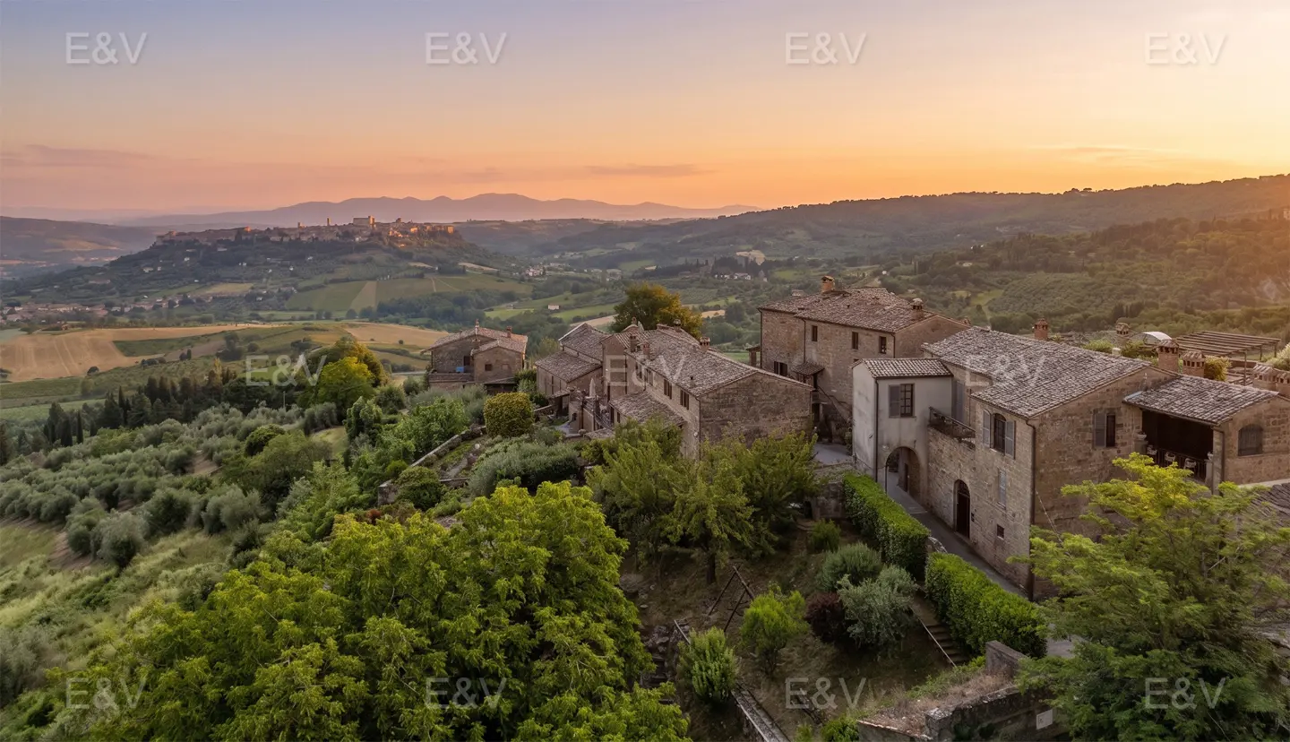 Aerial view of a stone village in the Tuscan countryside at sunset, surrounded by lush green hills and olive groves.