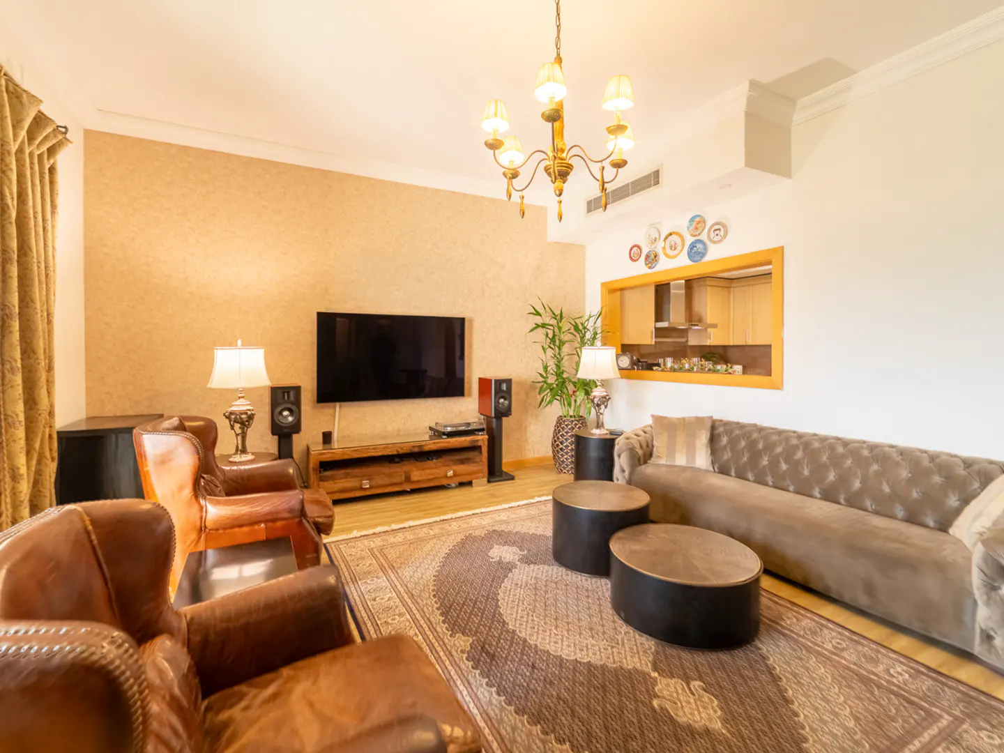 Living room with brown leather armchairs, a tufted sofa, two round coffee tables, and a wall-mounted TV on a wooden stand.