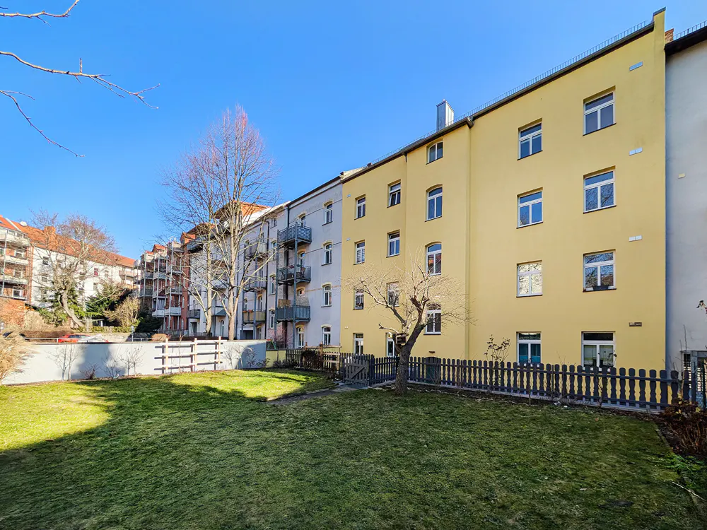 View of a yellow apartment building with a green lawn and a dark fence in front. Blue sky above.