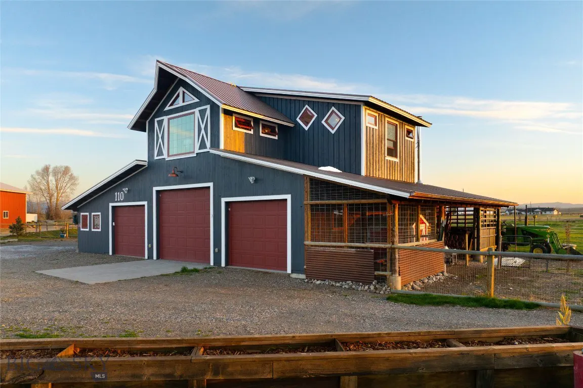 Two-story blue barn-style house with red roof and garage doors, set on a gravel lot with a green tractor in the background.