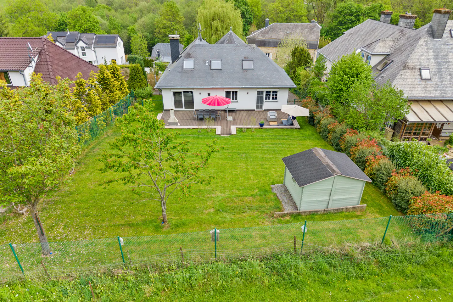 Aerial view of a white house with a gray roof, a green lawn, a wooden deck with furniture, and a small shed.