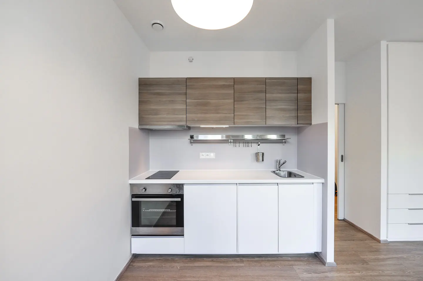 A modern kitchen with white cabinets, a stainless steel oven, and wood-look upper cabinets. A bright, round light fixture illuminates the space.