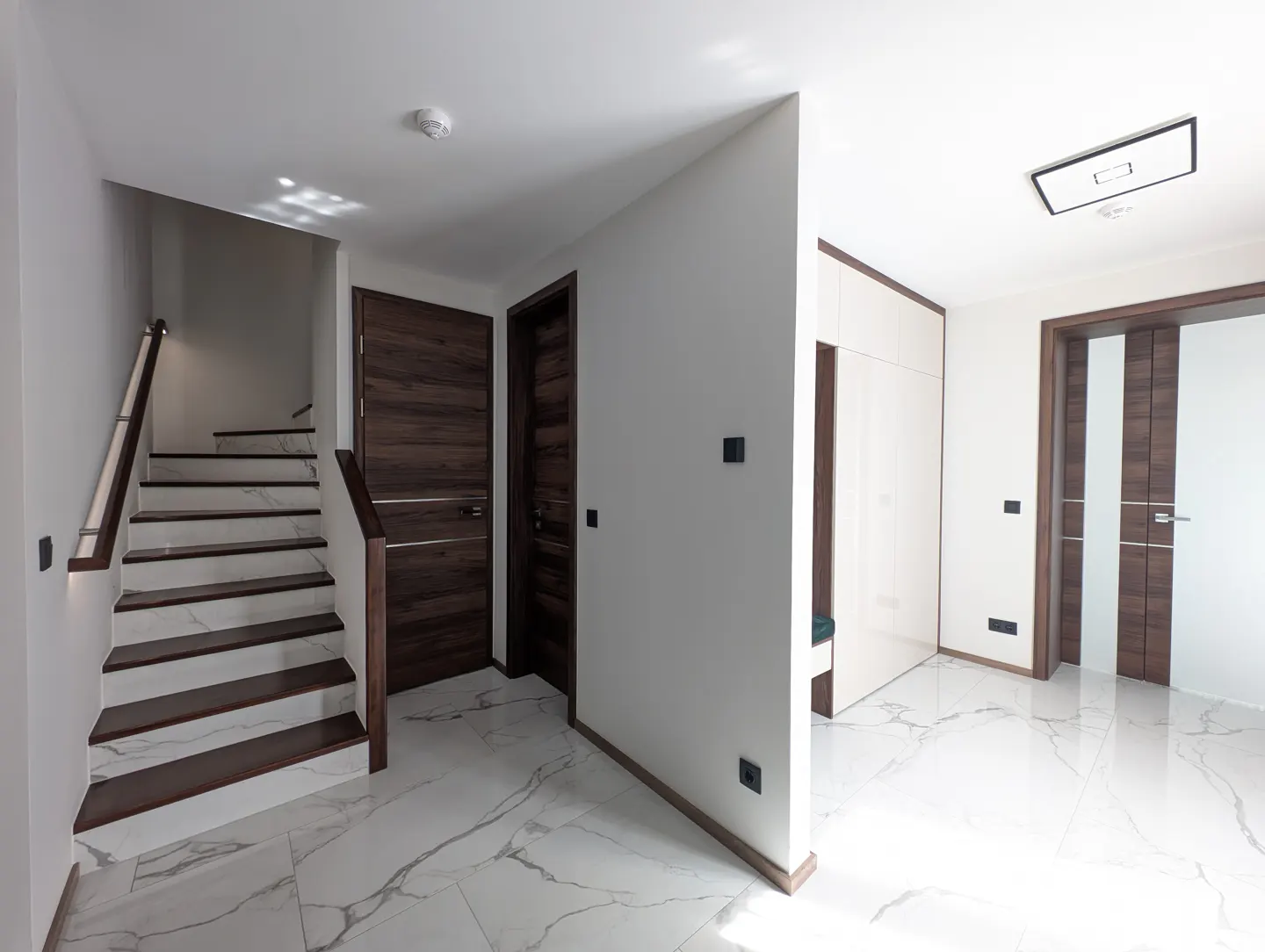 Bright foyer with marble floors, white walls, and dark wood accents. A staircase and doors are visible.