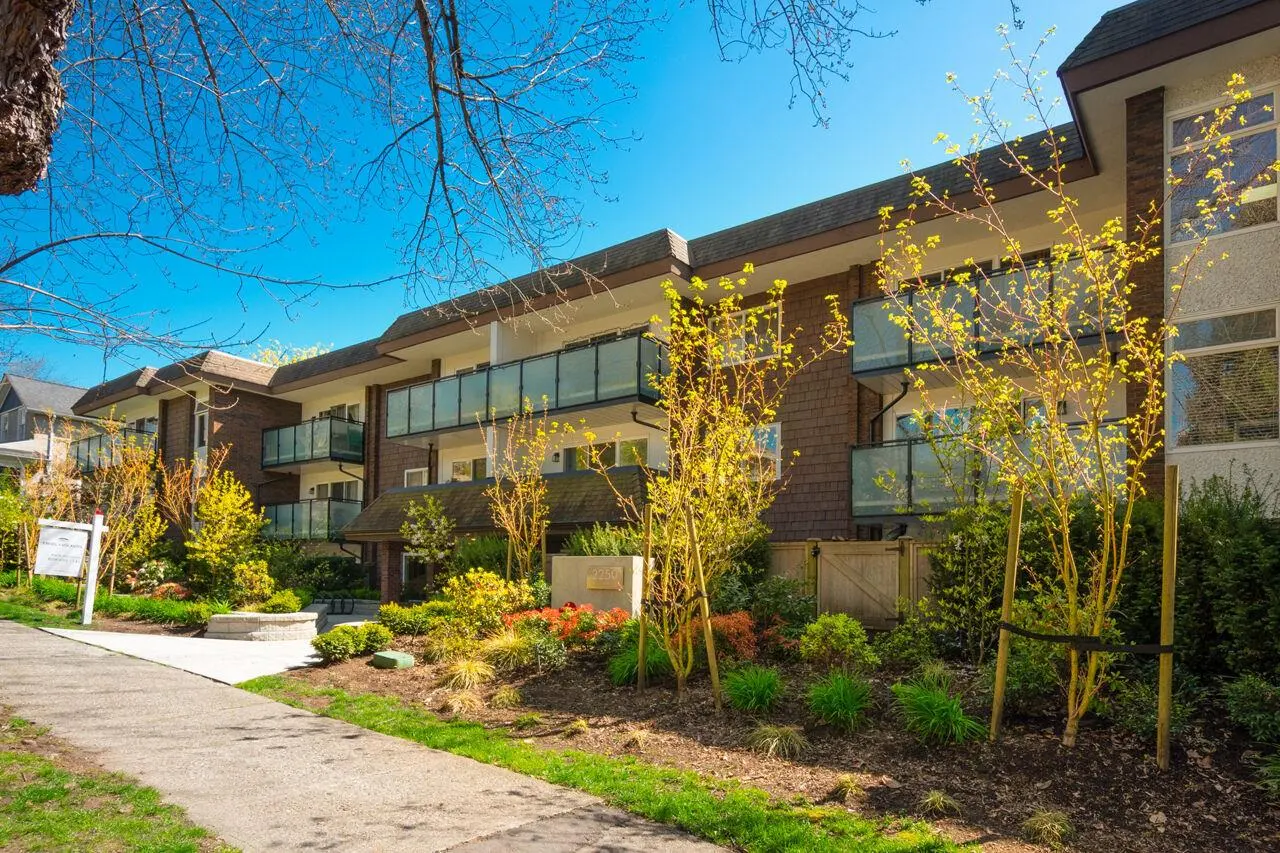 Exterior view of a brown two-story apartment building with glass balconies and manicured landscaping on a sunny day.