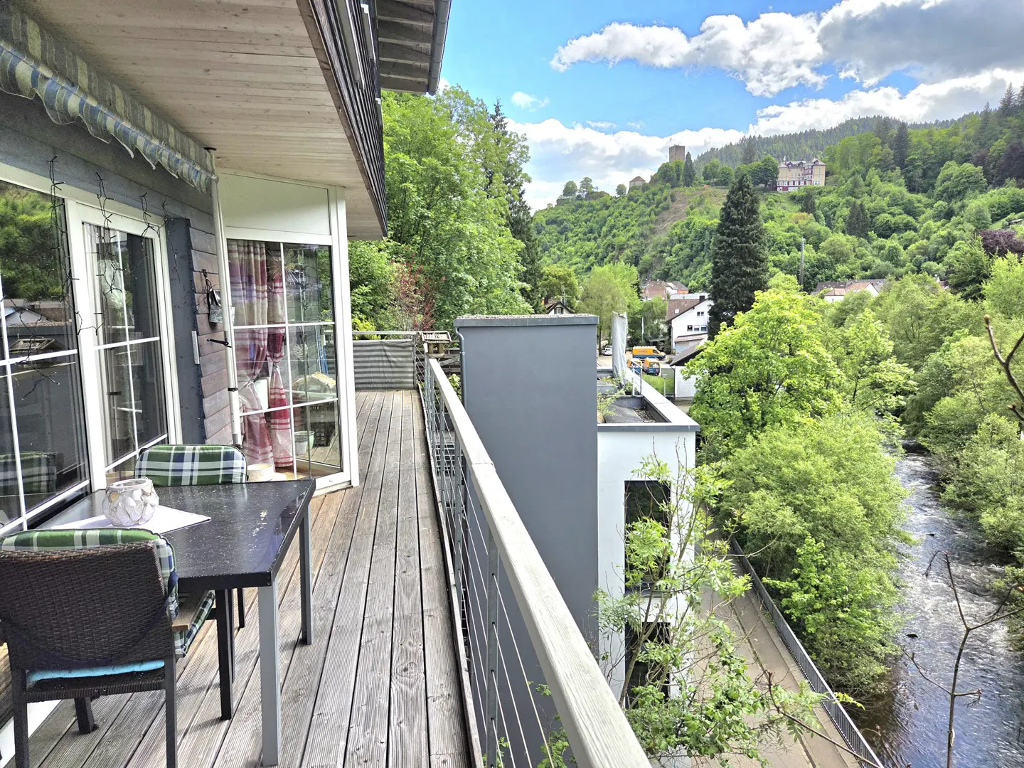 Balcony view with table, chairs, and a scenic backdrop of green hills, a river, and a castle under a blue sky.