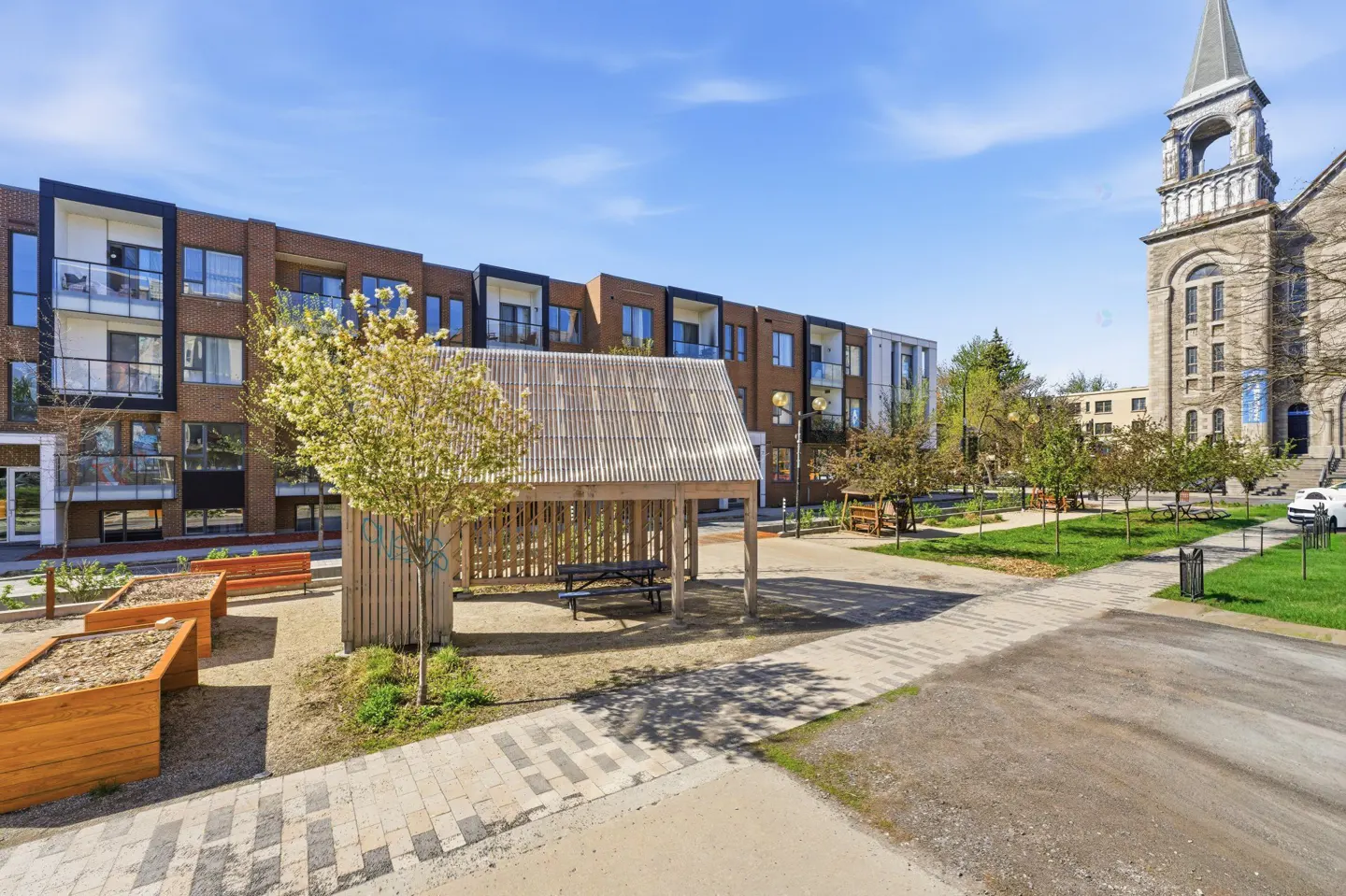 A modern brick apartment building overlooks a park with a wooden gazebo and benches, next to a stone church under a blue sky.