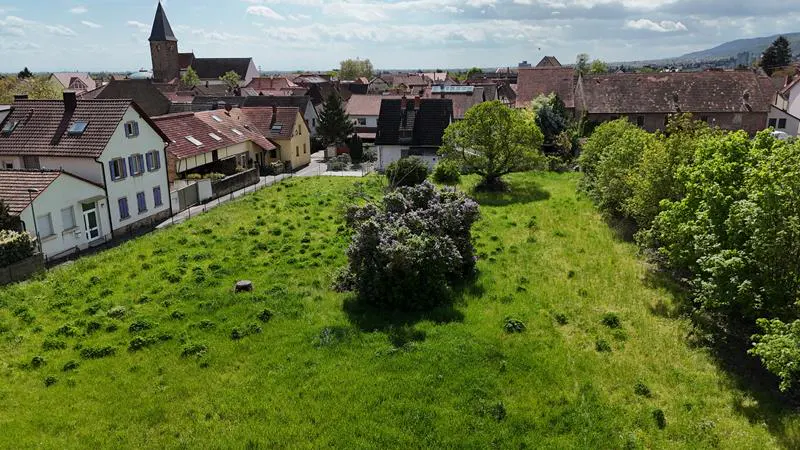 A vacant grassy lot in a European town, surrounded by houses with red tile roofs and a church steeple in the background.
