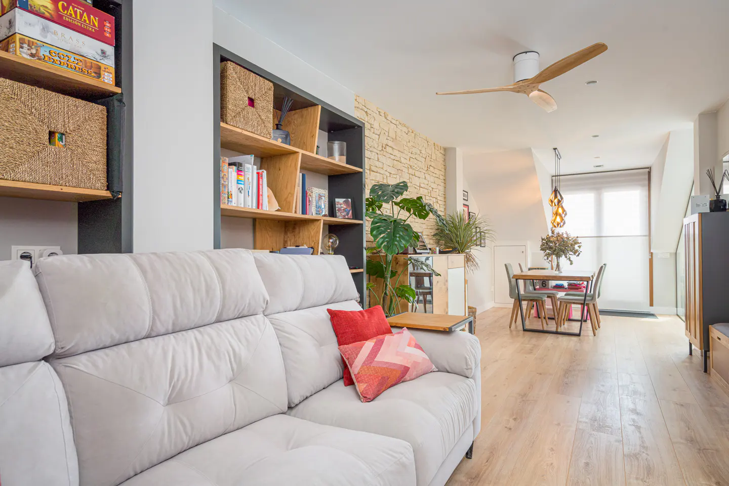 Bright living room with a white sofa, red pillows, and wooden shelves filled with books and baskets. A dining table and chairs are visible in the background.
