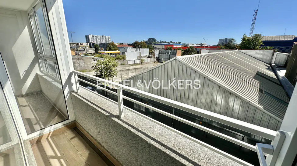 Balcony view with white railings overlooking a cityscape with buildings and a metal roof. The sky is blue.