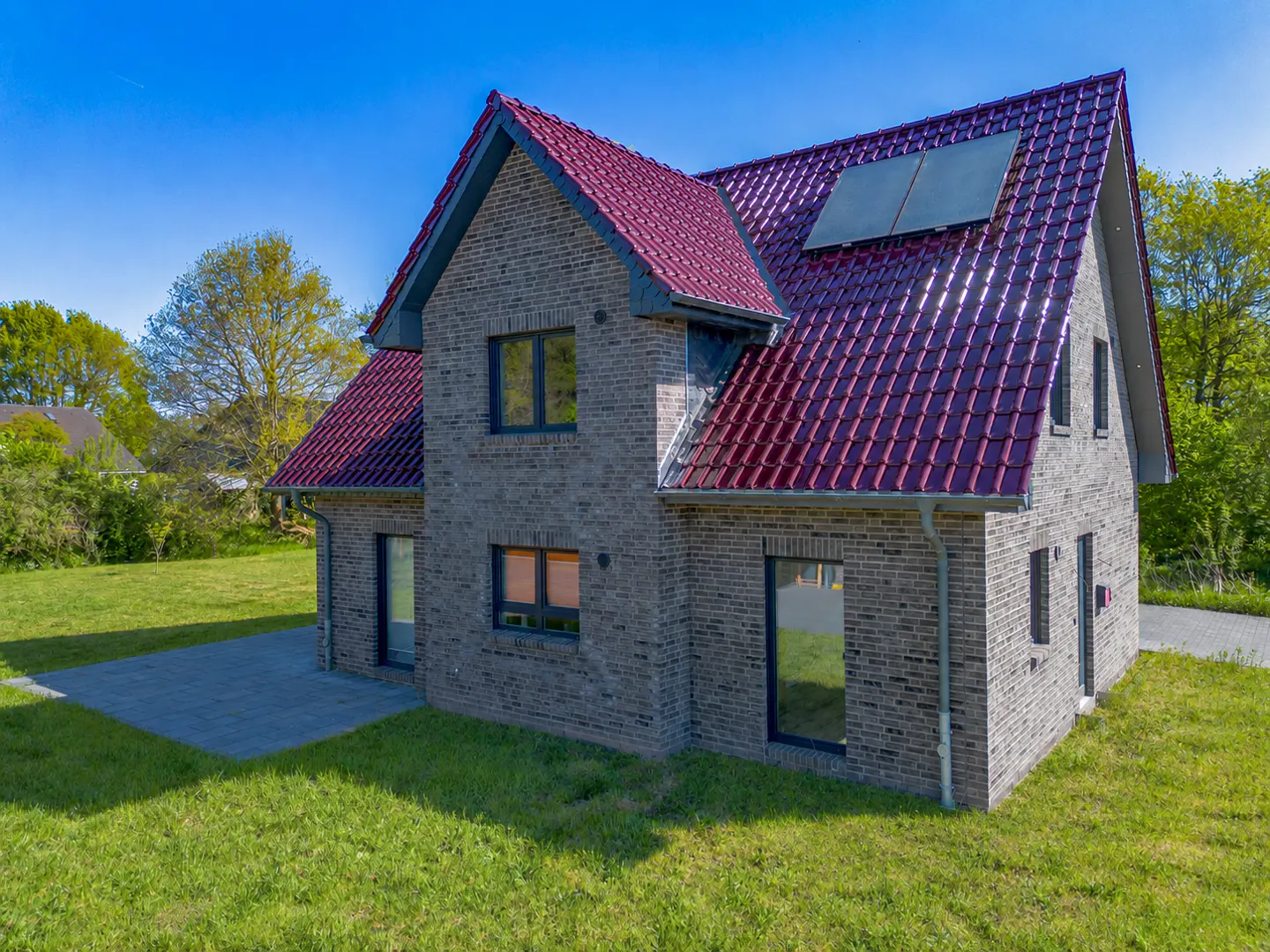 Two-story brick house with a red tile roof and solar panels, surrounded by green grass and trees under a blue sky.