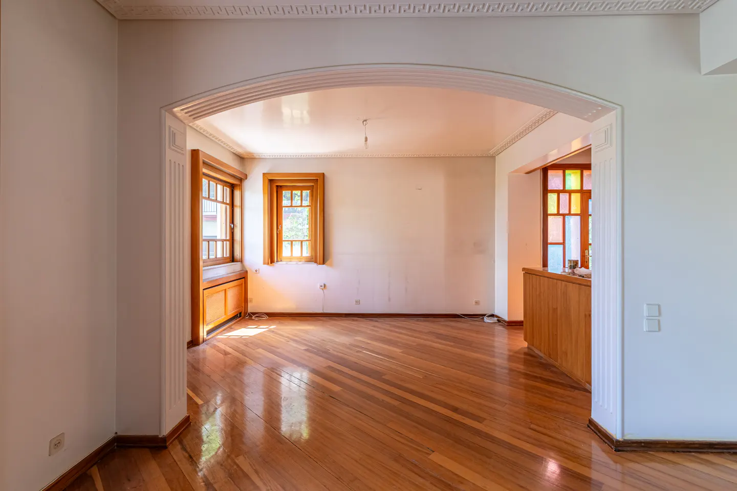 Empty room with hardwood floors, white walls, and arched doorway. Two wood-framed windows let in sunlight. Stained glass window in the background.