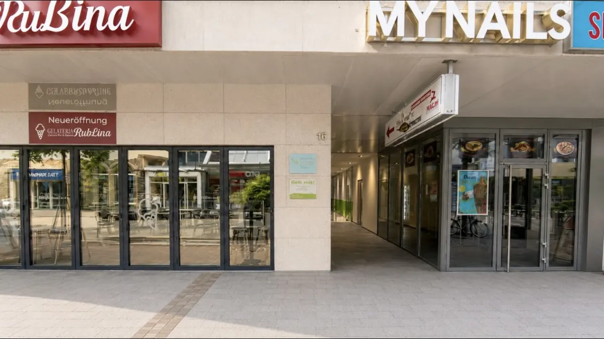 Exterior view of a shopping center with a Rublina Gelateria and a My Nails salon. The building is beige with glass doors and windows.