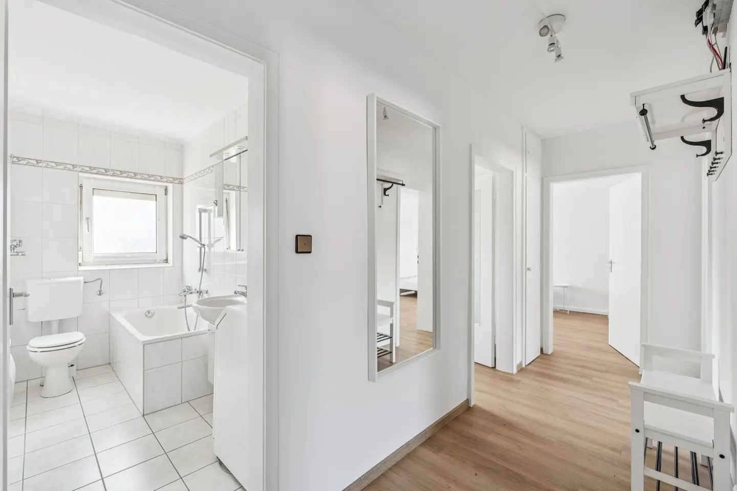 Bright hallway with wood floors, white walls, and doors. A bathroom is visible through an open doorway. A mirror and coat rack are on the wall.