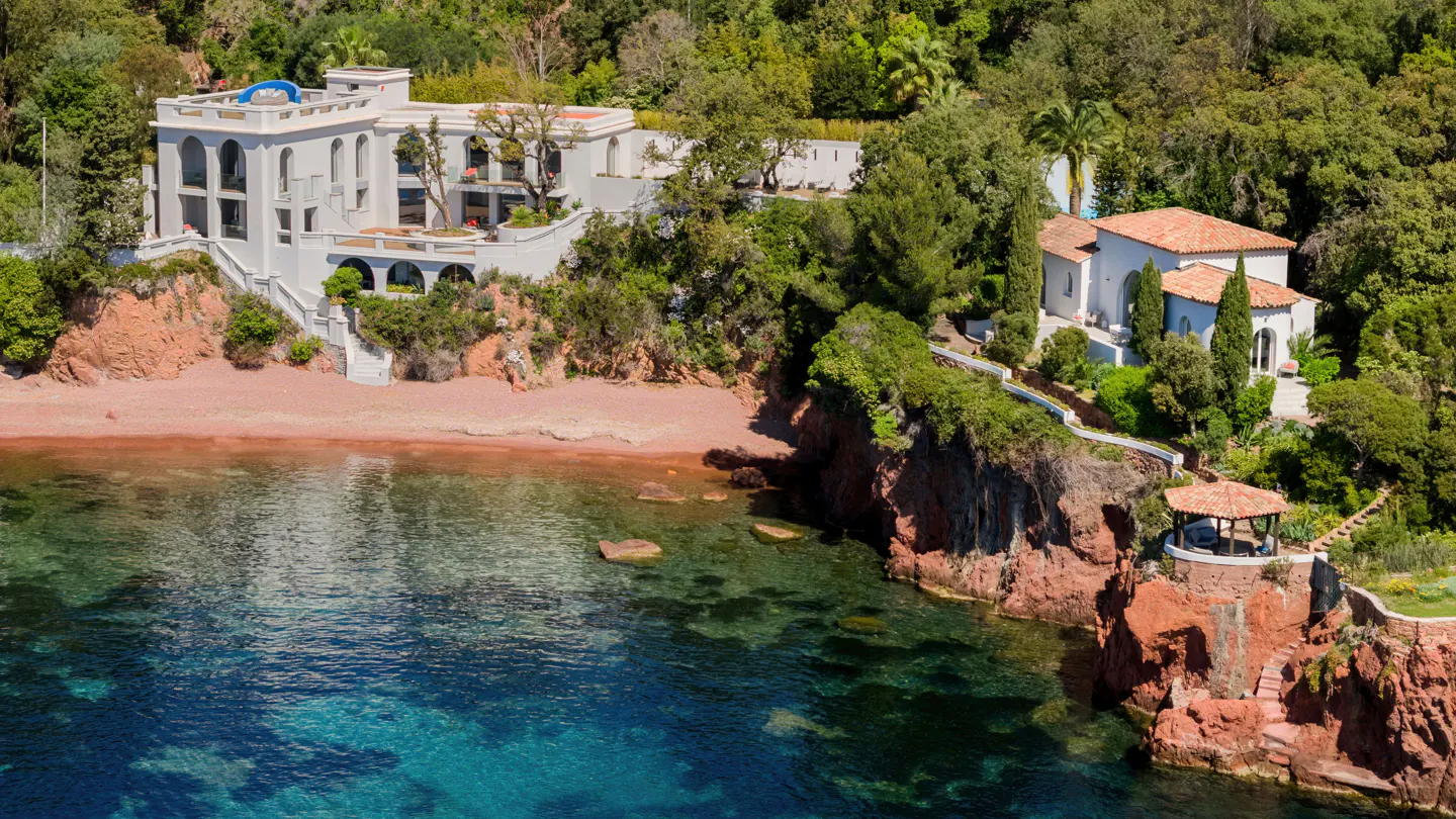 Aerial view of a white villa on a red cliff overlooking a beach and turquoise water, surrounded by green trees.