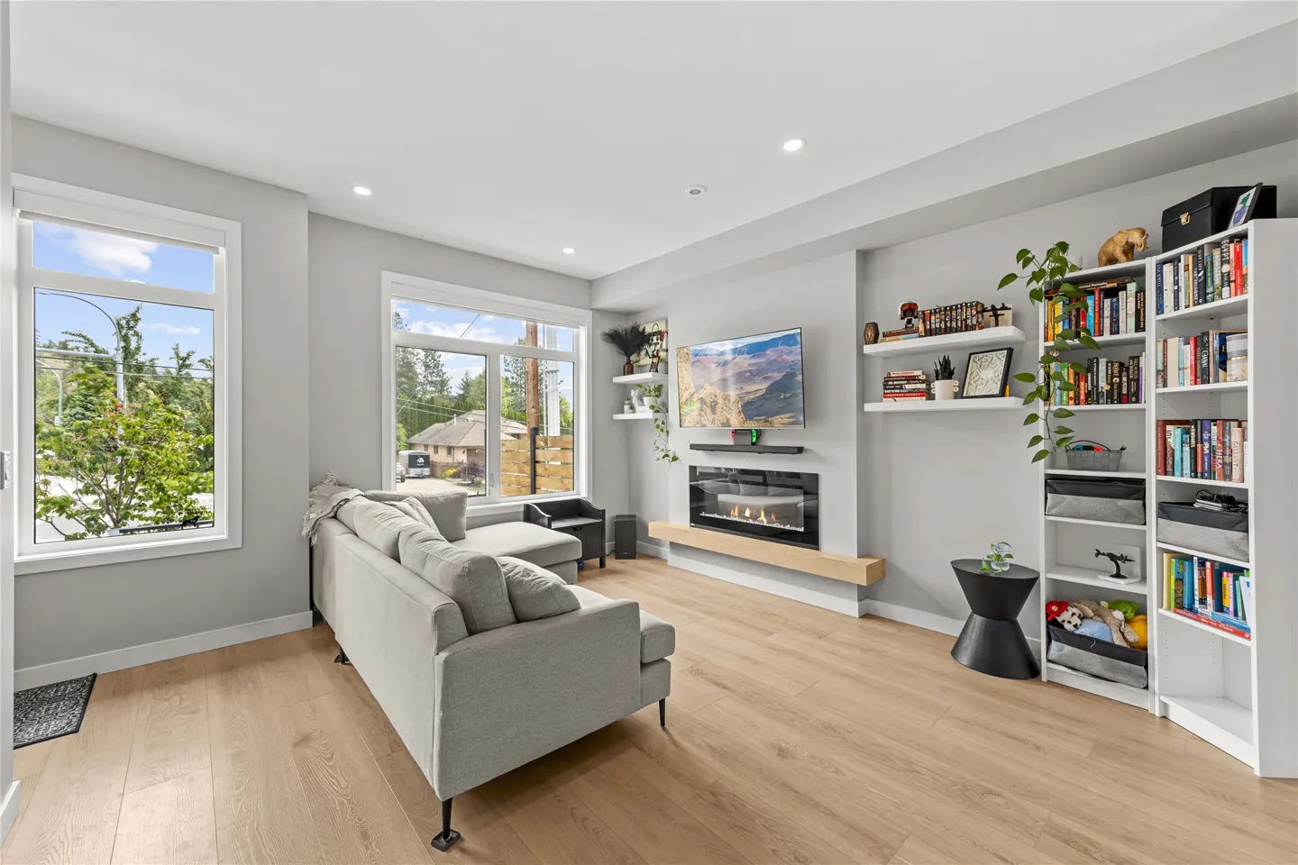 Bright living room with light wood floors, gray walls, and a gray sectional sofa. A fireplace and TV are mounted above. Bookshelves and windows add to the space.