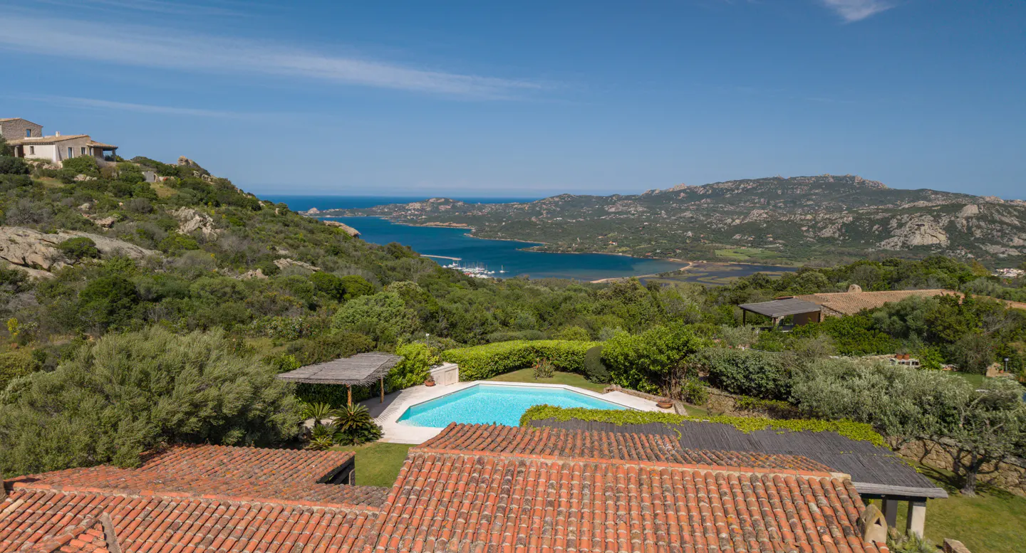 View of a blue pool and red tile roof, with a bay and mountains in the background.