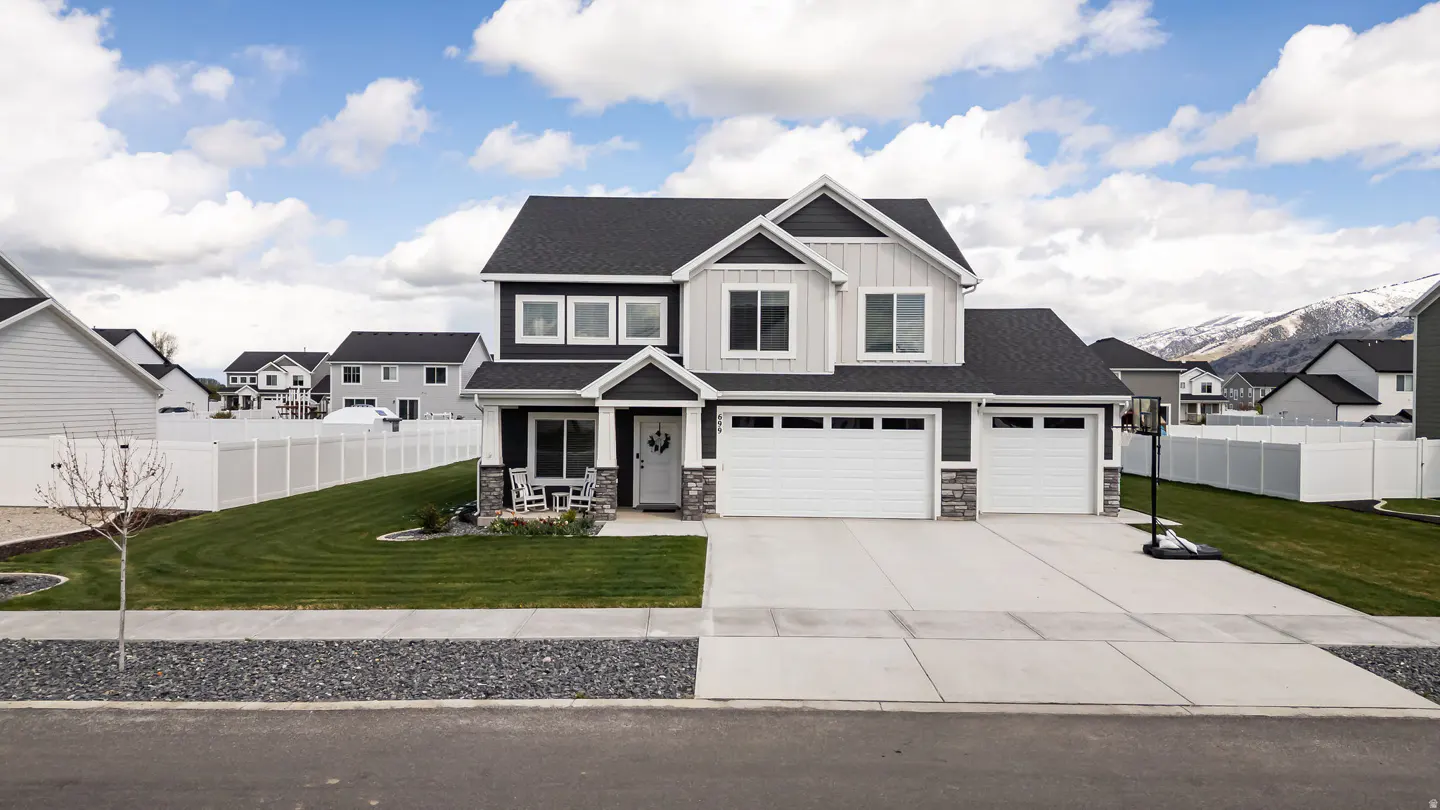 Two-story house with gray siding, white trim, and a black roof. A white fence surrounds the green lawn. Mountains are visible in the background.