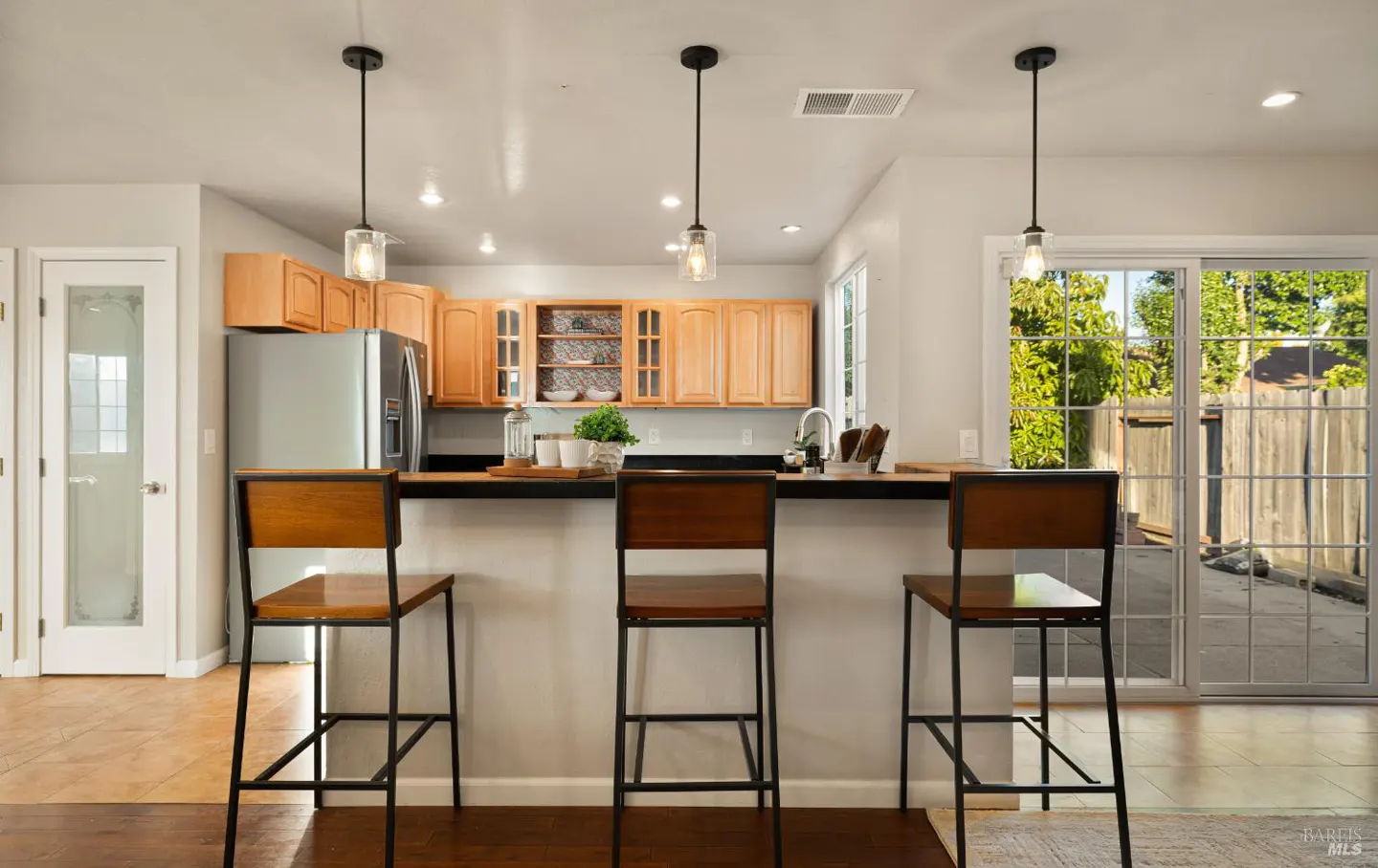 A kitchen with wood cabinets, stainless steel appliances, and three bar stools at a black countertop. Three pendant lights hang above. Sliding glass doors lead to a backyard.
