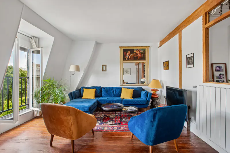 Bright living room with a blue sectional sofa, orange and blue armchairs, a patterned rug, and a balcony with greenery.