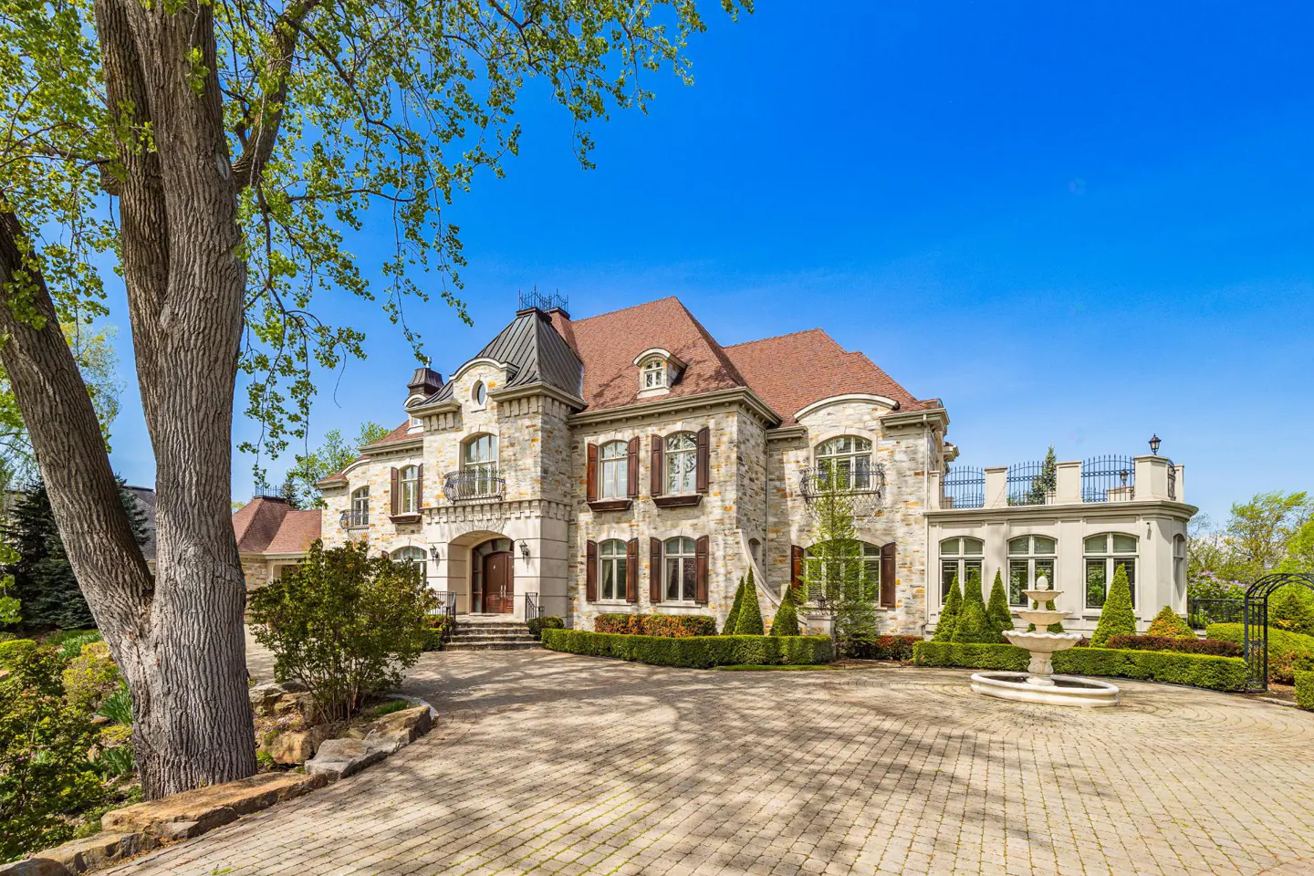 Exterior view of a large stone house with a brown roof, brown shutters, and a circular driveway with a fountain.