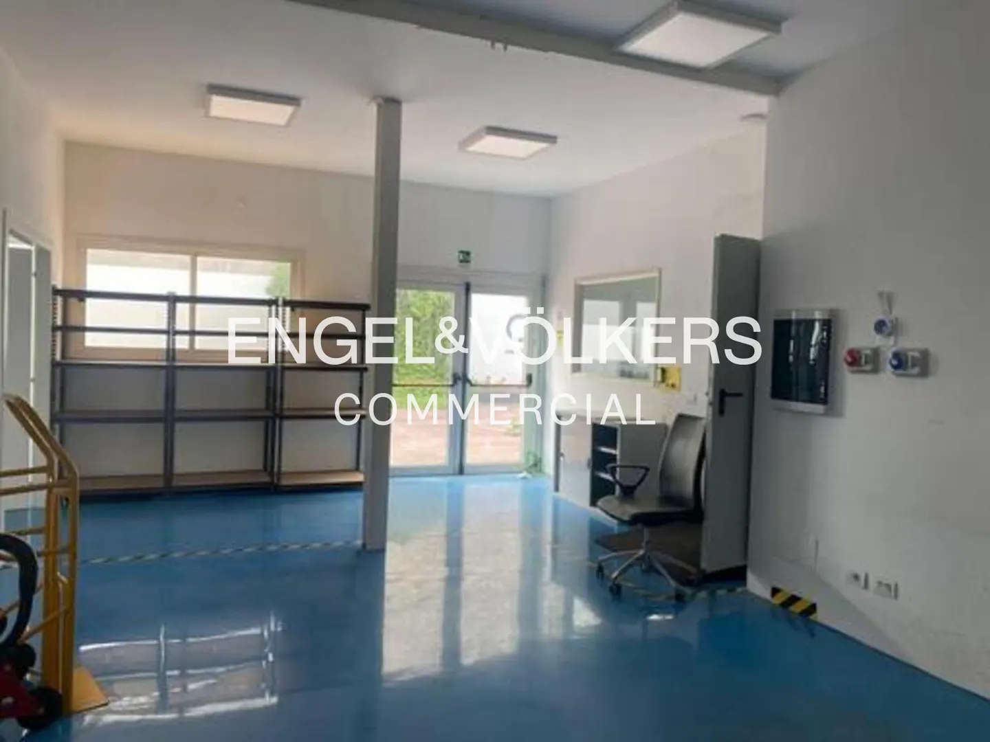 Bright, empty commercial space with blue floor, white walls, shelves, and a rolling chair. A hand truck is in the foreground.