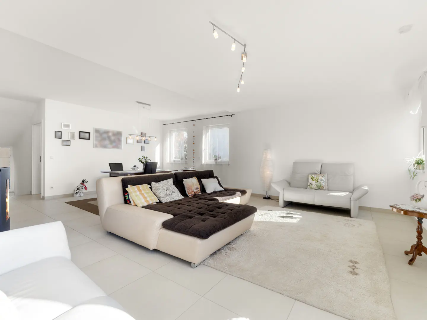 Bright, open living room with white walls, tile floors, and a large brown and beige sectional sofa. A white rug covers the floor.
