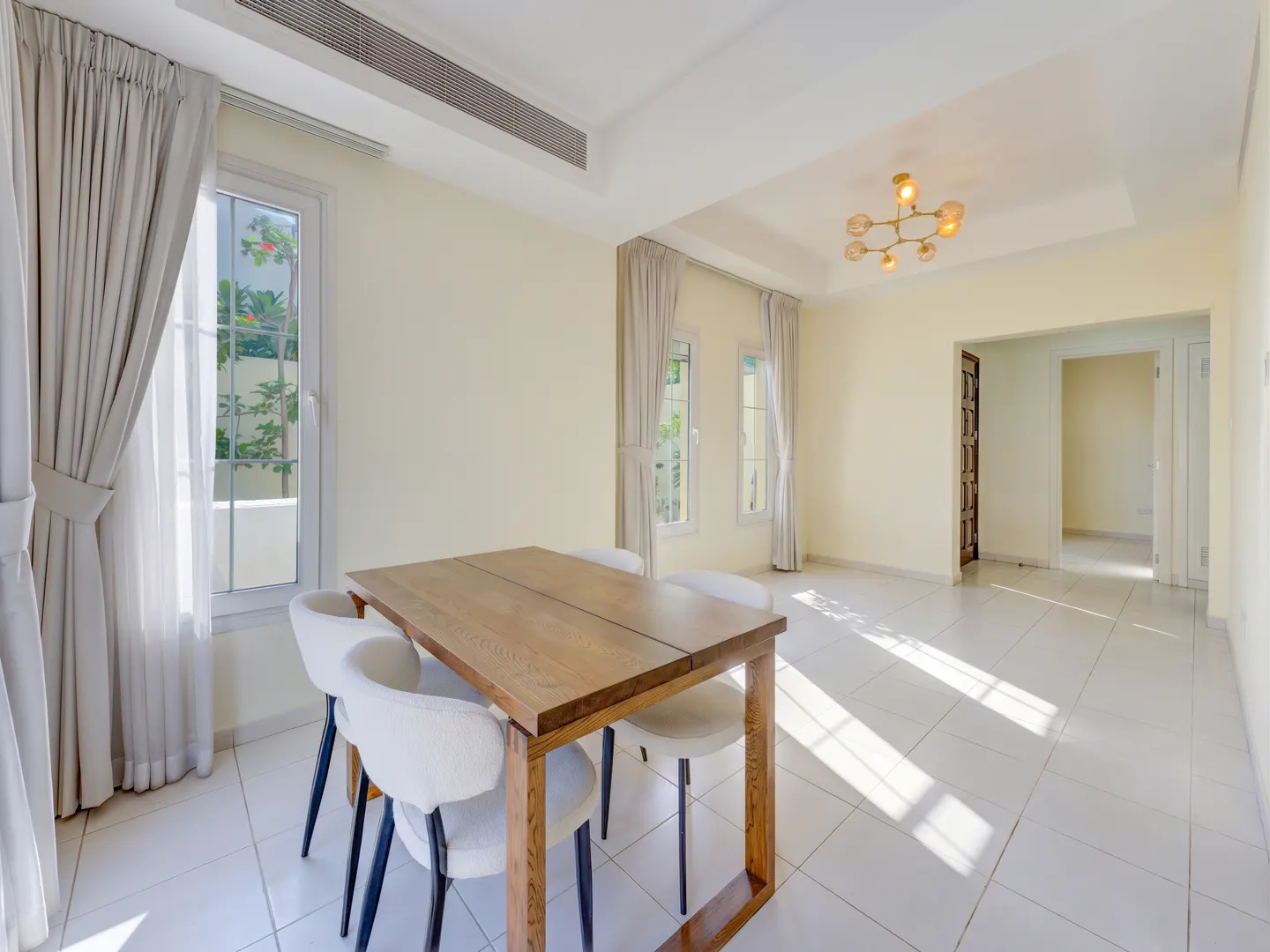 Bright dining room with a wooden table, four white chairs, light-colored walls, tile floors, and large windows with curtains.