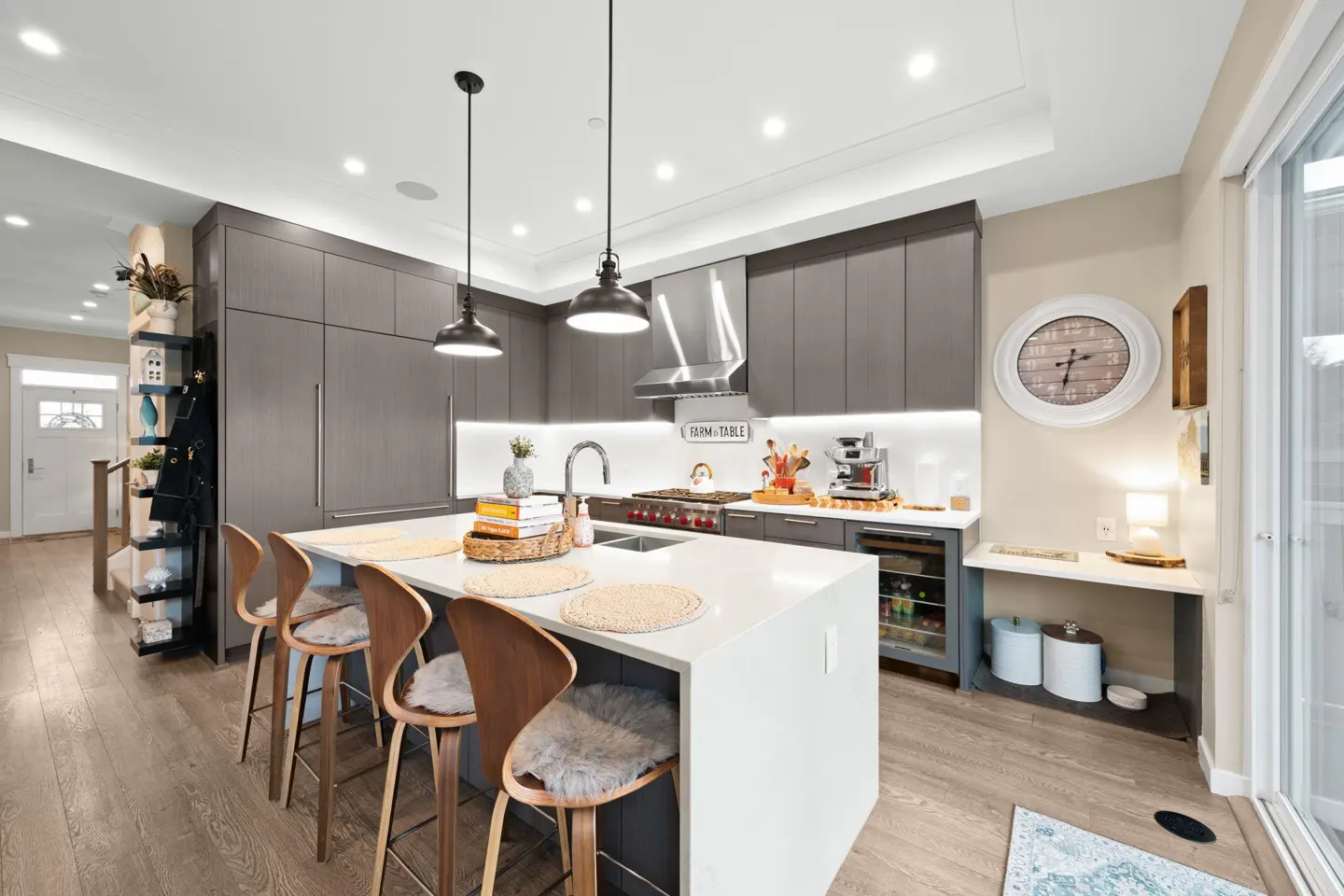 A modern kitchen with gray cabinets, a white island with four stools, and stainless steel appliances. Pendant lights hang above the island.