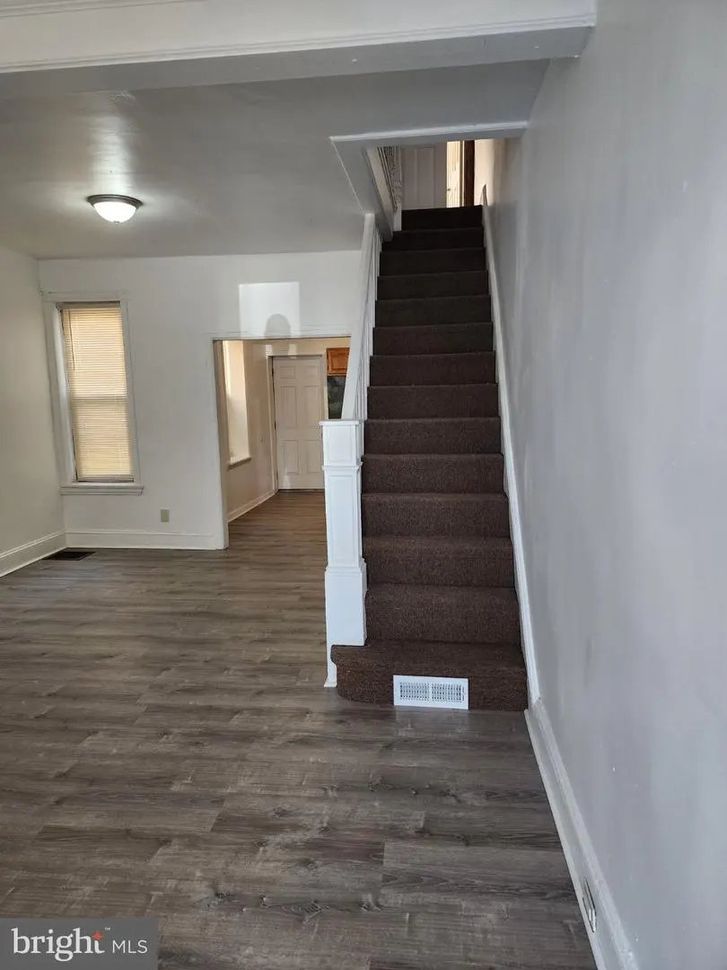 Interior view of a home with gray wood floors, white walls, and carpeted stairs leading up. A doorway leads to another room.