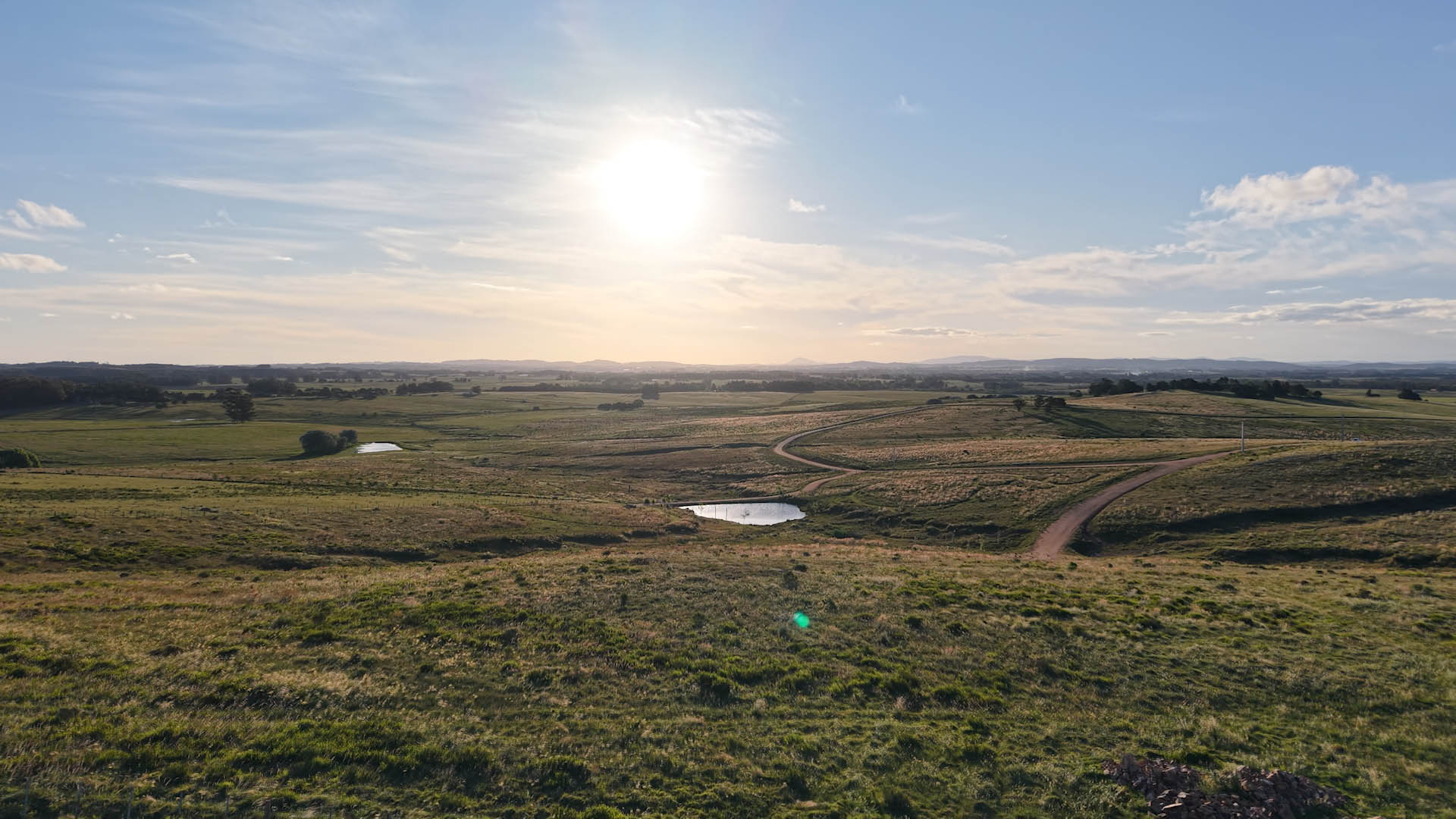 Aerial view of a vast, rolling grassy landscape with small ponds and dirt paths under a bright, sunlit sky.