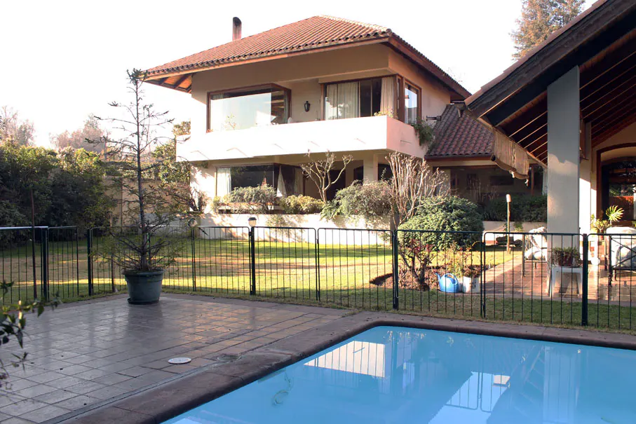 Two-story beige house with a red tile roof, viewed from the pool area with a black metal fence.