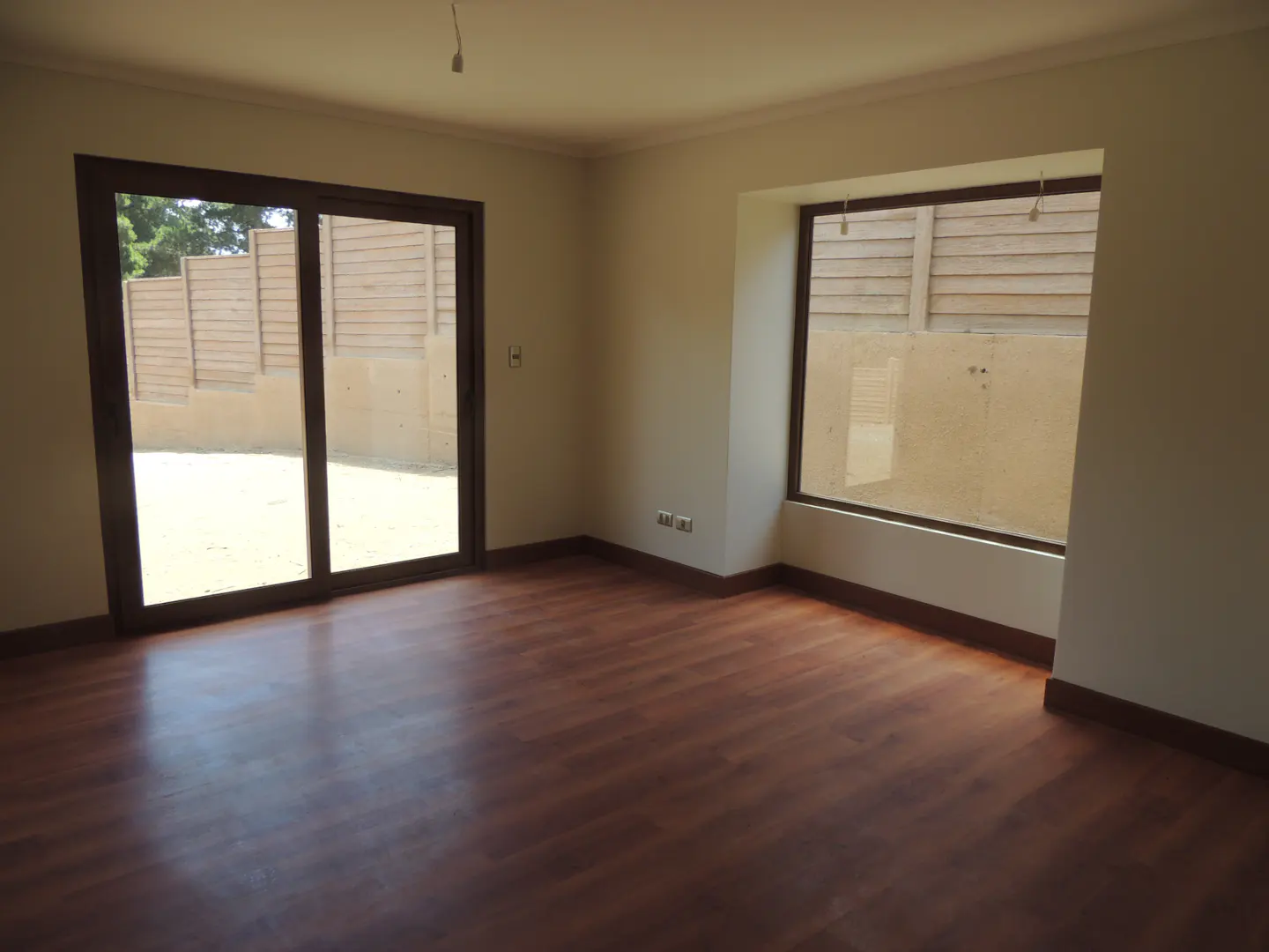 Empty room with brown wood floor, white walls, and large windows. A sliding glass door leads to a fenced backyard.