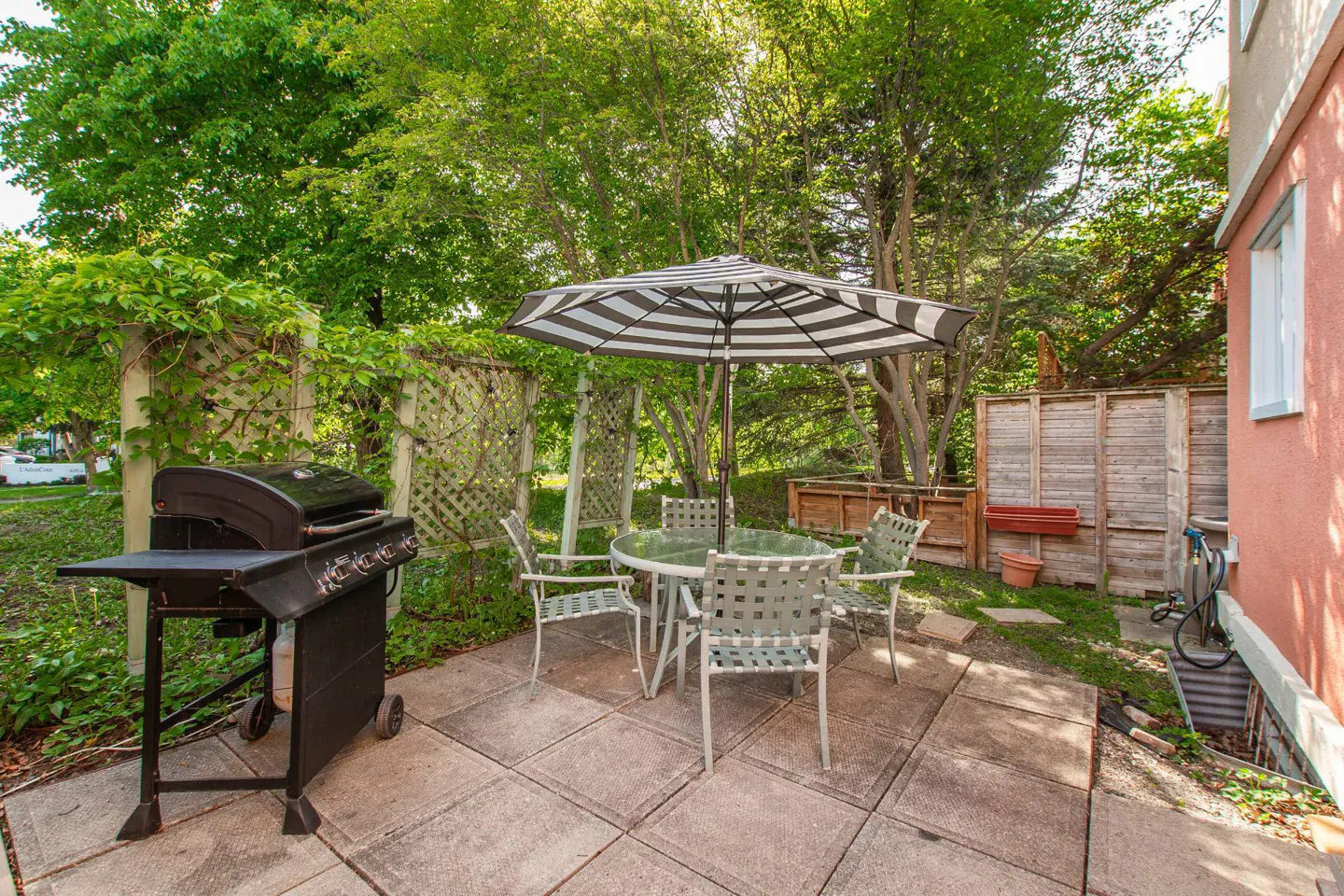 Backyard patio with a black grill, a glass table, four chairs, and a black-and-white striped umbrella under lush trees.